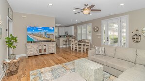 A spacious living area with a large flat-screen TV, a ceiling fan, and a dining area with a kitchen in the background.