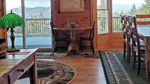 A wooden cabin interior with a dining table, chairs, a map on the wall, and large windows overlooking a scenic view.