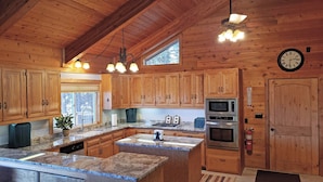 A wooden cabin kitchen with granite countertops, wooden cabinets, a microwave, oven, and a clock on the wall.