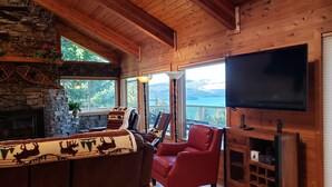 A cozy cabin interior with a stone fireplace, wooden walls, a large TV, and a view of the outdoors.