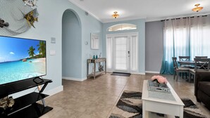 A living room with a TV displaying a beach scene, a dining area with a table and chairs, and a hallway with a door and a small table with decorative items. - Stylish & Bright, Mins to Downtown, Pool, Parking (North Port)