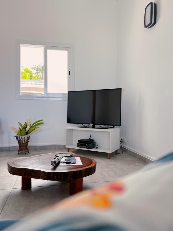 A modern living room with a flat-screen TV on a white stand, a wooden coffee table with remote controls, and a potted plant.