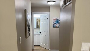 A hallway with wooden flooring, beige walls, a ceiling light, a vent, and a door leading to a bathroom. - Wilson, Two Bedroom House in Historic Loveland (Loveland)