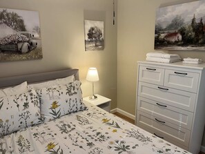 Et soveværelse med en seng, et natbord med lampe, en kommode og to billeder på væggen.