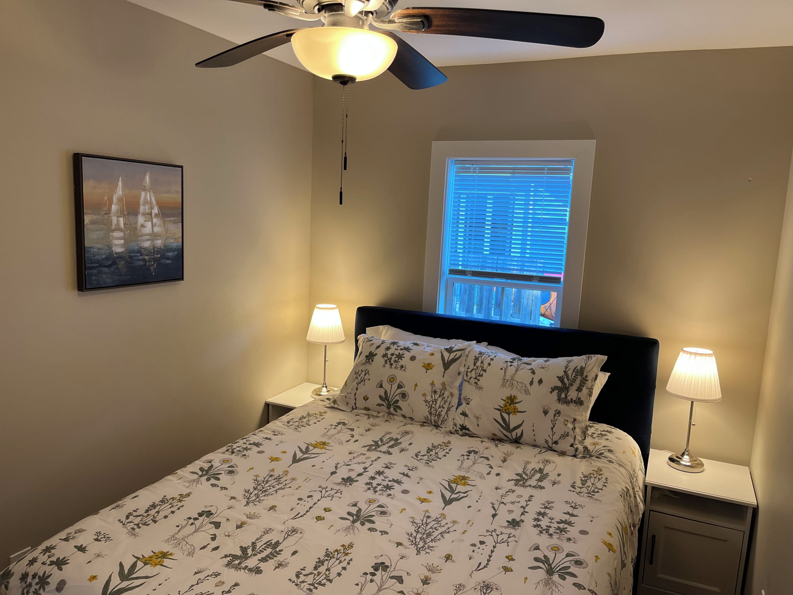 Una camera da letto con un letto, due lampade sui comodini, una ventola a soffitto, una finestra con persiane e un quadro appeso al muro.