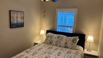 Una camera da letto con un letto, due lampade sui comodini, una ventola a soffitto, una finestra con persiane e un quadro appeso al muro.