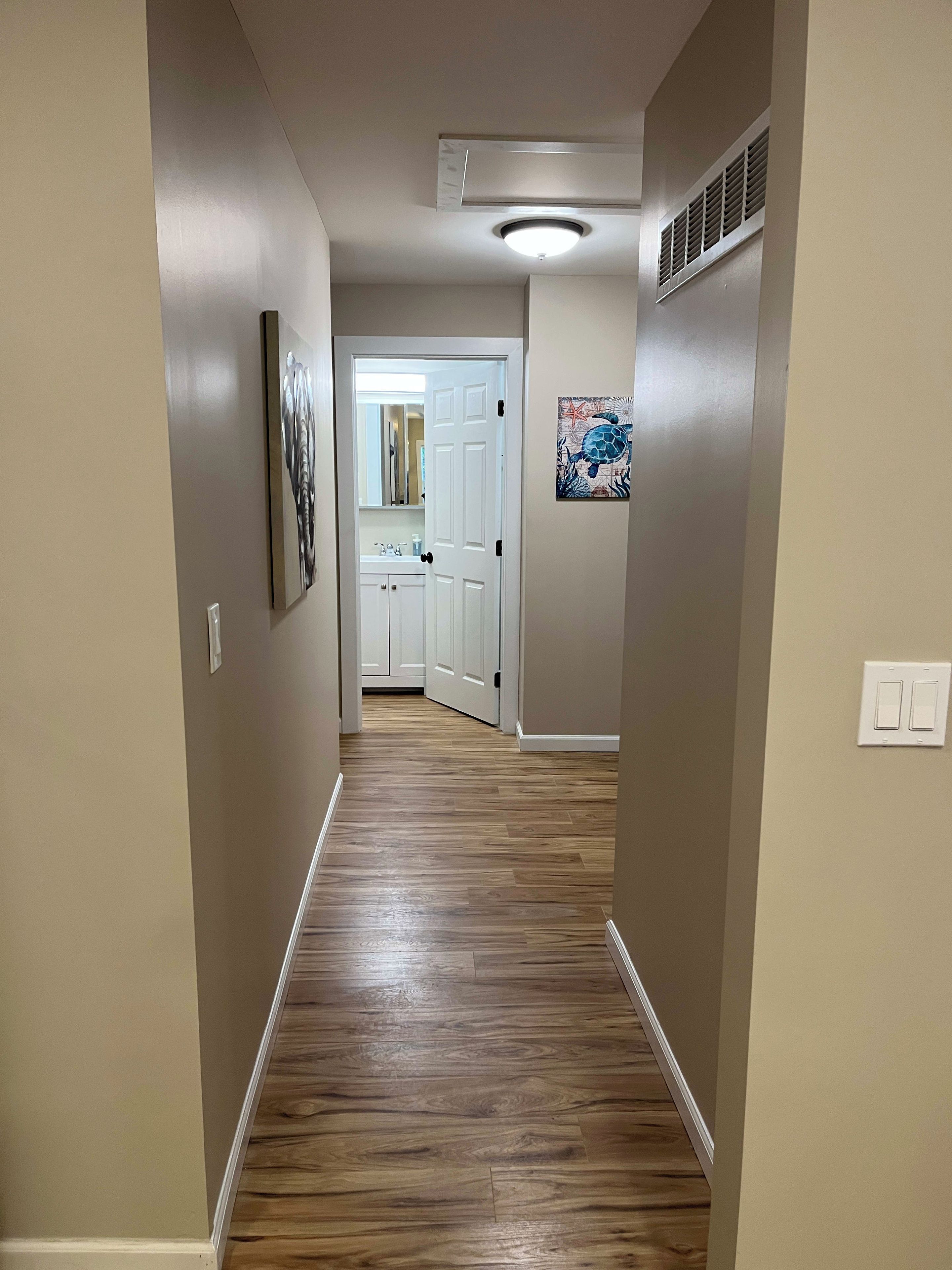 A hallway with wooden flooring, beige walls, a ceiling light, a vent, and a door leading to a bathroom.