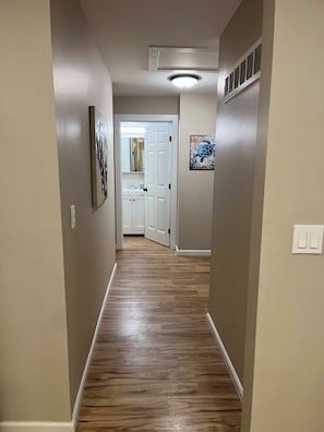 A hallway with wooden flooring, beige walls, a ceiling light, a vent, and a door leading to a bathroom. - Wilson, Two Bedroom House in Historic Loveland (Loveland)