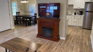 A modern living room with a fireplace, a TV mounted on the wall, a dining area, and a kitchen with white cabinets and stainless steel appliances. - Wilson, Two Bedroom House in Historic Loveland (Loveland)