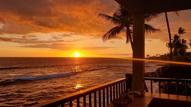 Vista al atardecer desde un balcón con barandilla, palmeras y olas del océano.