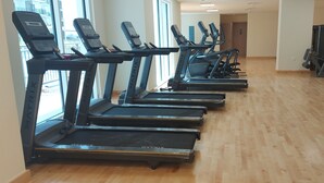 Een sportschool met meerdere loopbanden op een rij voor een raam, een houten vloer en diverse fitnessapparaten.