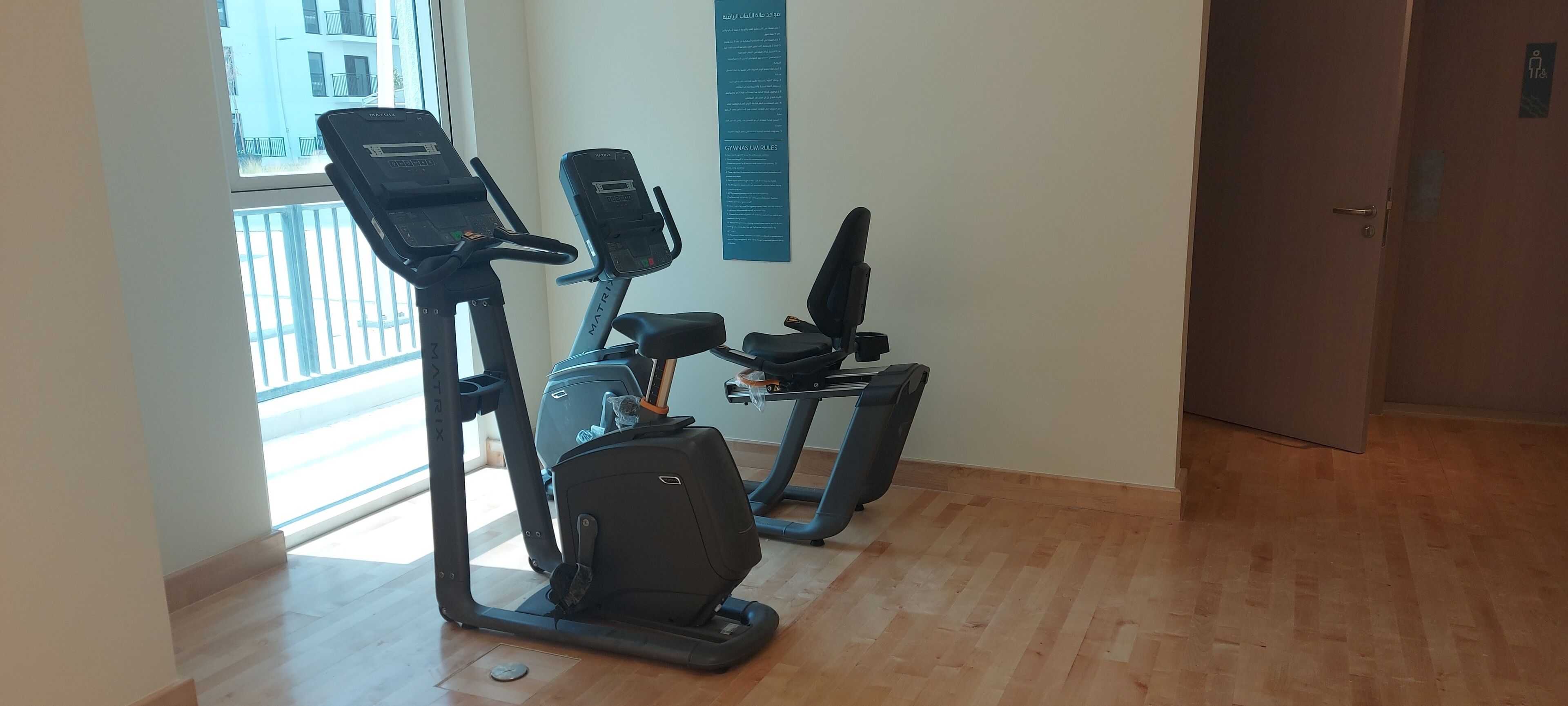 Een sportschool met loopbanden en een elliptische machine. Er is een raam met uitzicht op gebouwen.