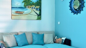 A living room with a sofa, pillows, a glass coffee table, a painting of a beach scene, and a decorative mirror. - Studio Indépendant Devant Piscine et BBQ à Orient Bay à 5 Minutes de la Plage ! (saint martin)