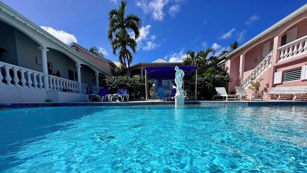 A clear blue pool with lounge chairs and a statue in the center, surrounded by palm trees and a building with a balcony. - Studio Indépendant Devant Piscine et BBQ à Orient Bay à 5 Minutes de la Plage ! (saint martin)