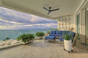 A balcony with a view of the sea, a ceiling fan, a blue sofa, and potted plants.