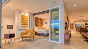 A modern hotel room with a large bed, a seating area with a chair, a desk, and a glass partition leading to a balcony.