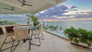 A balcony with a view of the sunset over the sea, wicker furniture, ceiling fans, and potted plants.