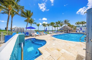 A pool area with lounge chairs, palm trees, and a thatched-roof structure. - Gorgeous Sunny Isles Beach Condo w/Balcony an Ocean View & Direct Beach Access! (Sunny Isles Beach)