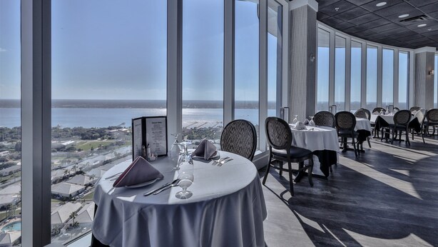 Un coin repas avec une table dressée pour deux personnes, de grandes fenêtres offrant une vue sur la mer et un plafond haut doté d’un éclairage encastré.