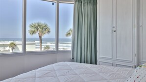 Une chambre avec une grande fenêtre donnant sur la plage, un lit blanc et une armoire.