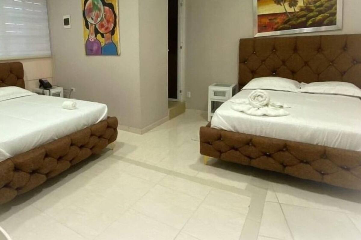 A hotel room with two beds, a small bedside table, a wall painting, and a window with blinds.