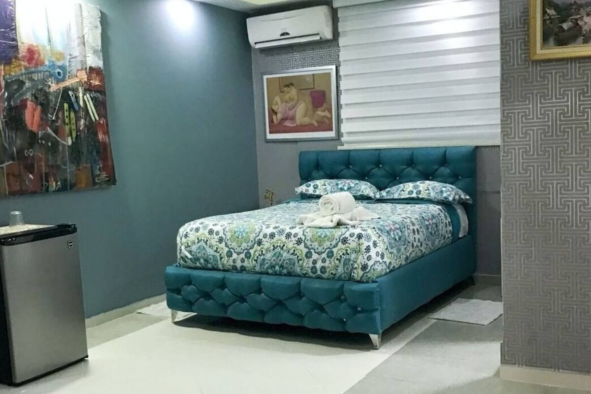 A bedroom with a teal upholstered bed, patterned bedding, a small refrigerator, and a wall-mounted air conditioner.