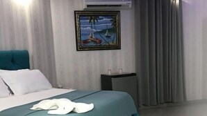 A neatly made bed with a teal bedspread and white pillows, a small bedside table with two cups, and a framed painting of a sailboat on the wall.