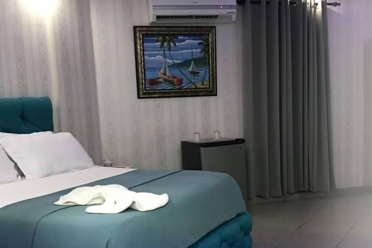 A neatly made bed with a teal bedspread and white pillows, a small bedside table with two cups, and a framed painting of a sailboat on the wall.