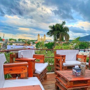 A wooden deck with outdoor seating, a view of a historic building with two towers, and a palm tree in the background. - Dinasty Hotel (Puerto Plata)