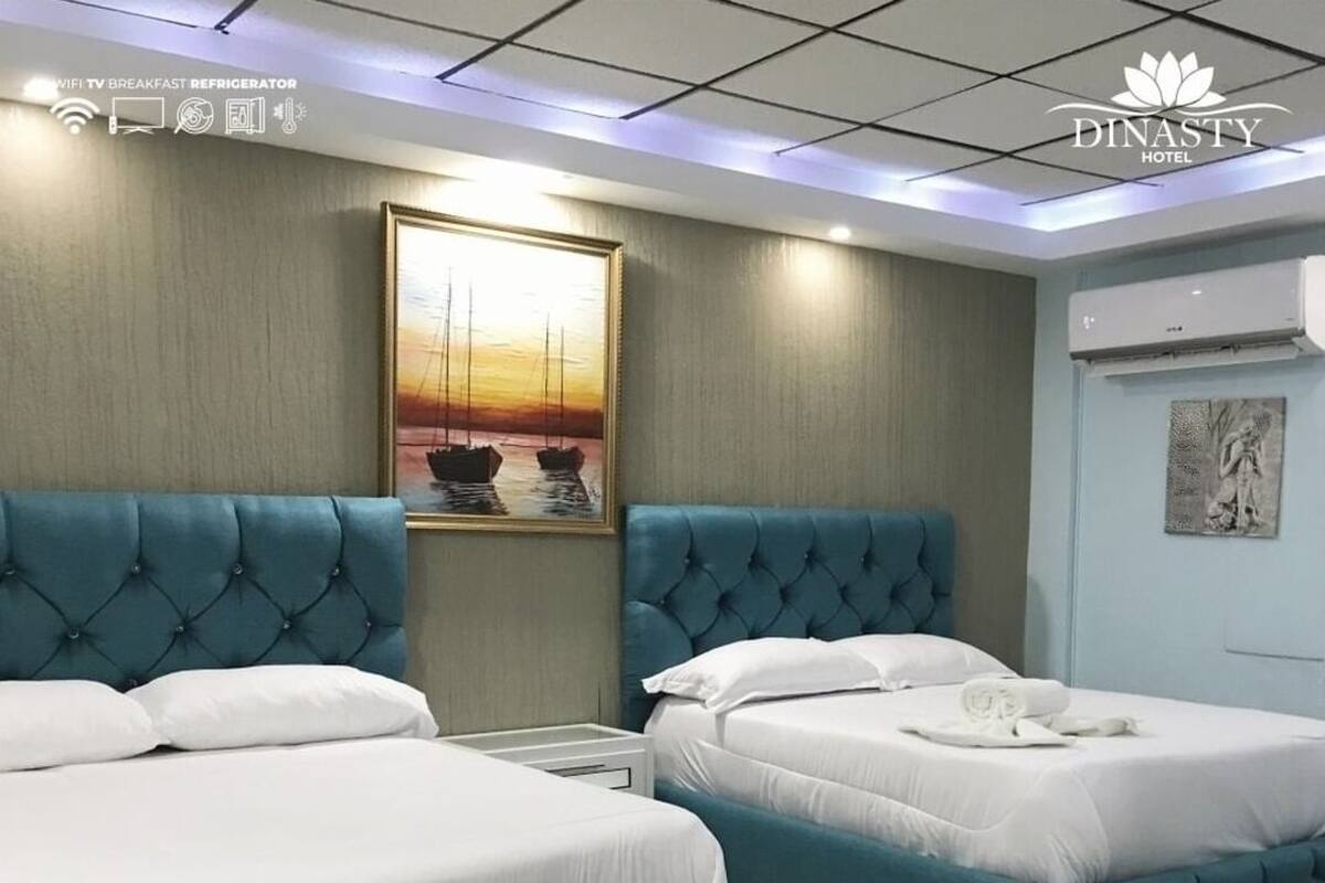 A hotel room with two beds, a painting of a sunset, and a sign indicating WIFI, TV, breakfast, and refrigerator amenities.