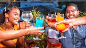 A group of people toasting with colorful drinks at an outdoor setting with a cityscape in the background.