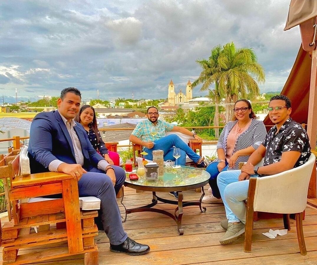 A group of people seated on outdoor furniture on a wooden deck with a glass table, overlooking a scenic view with a church and palm trees.