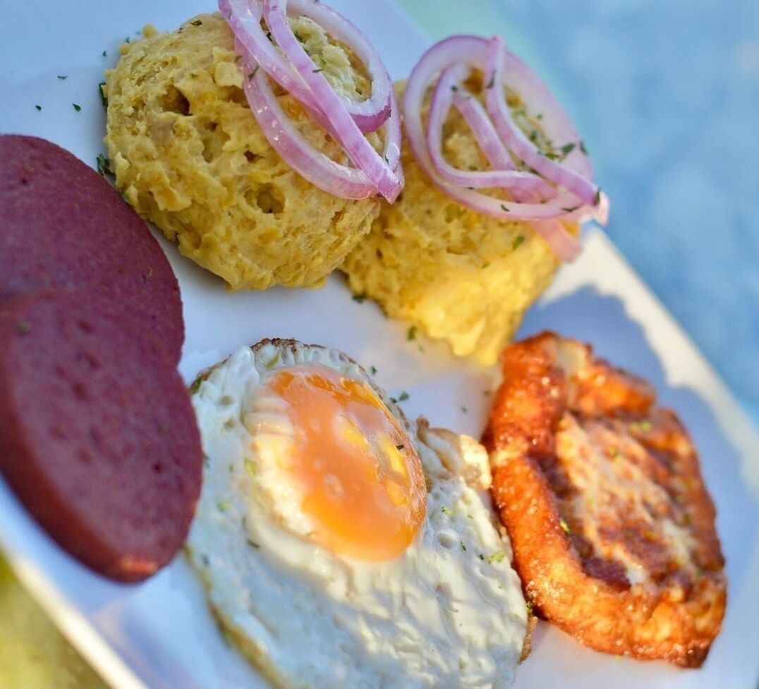 Une assiette contenant un œuf au plat, une tranche de saucisse, un morceau de banane plantain frite et une petite portion de nourriture garnie d’oignons rouges en tranches.