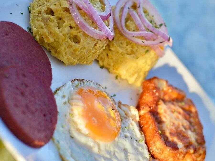 Un plato con un huevo frito, una rebanada de salchicha, un trozo de plátano frito y una pequeña porción de comida cubierta con rodajas de cebolla roja.