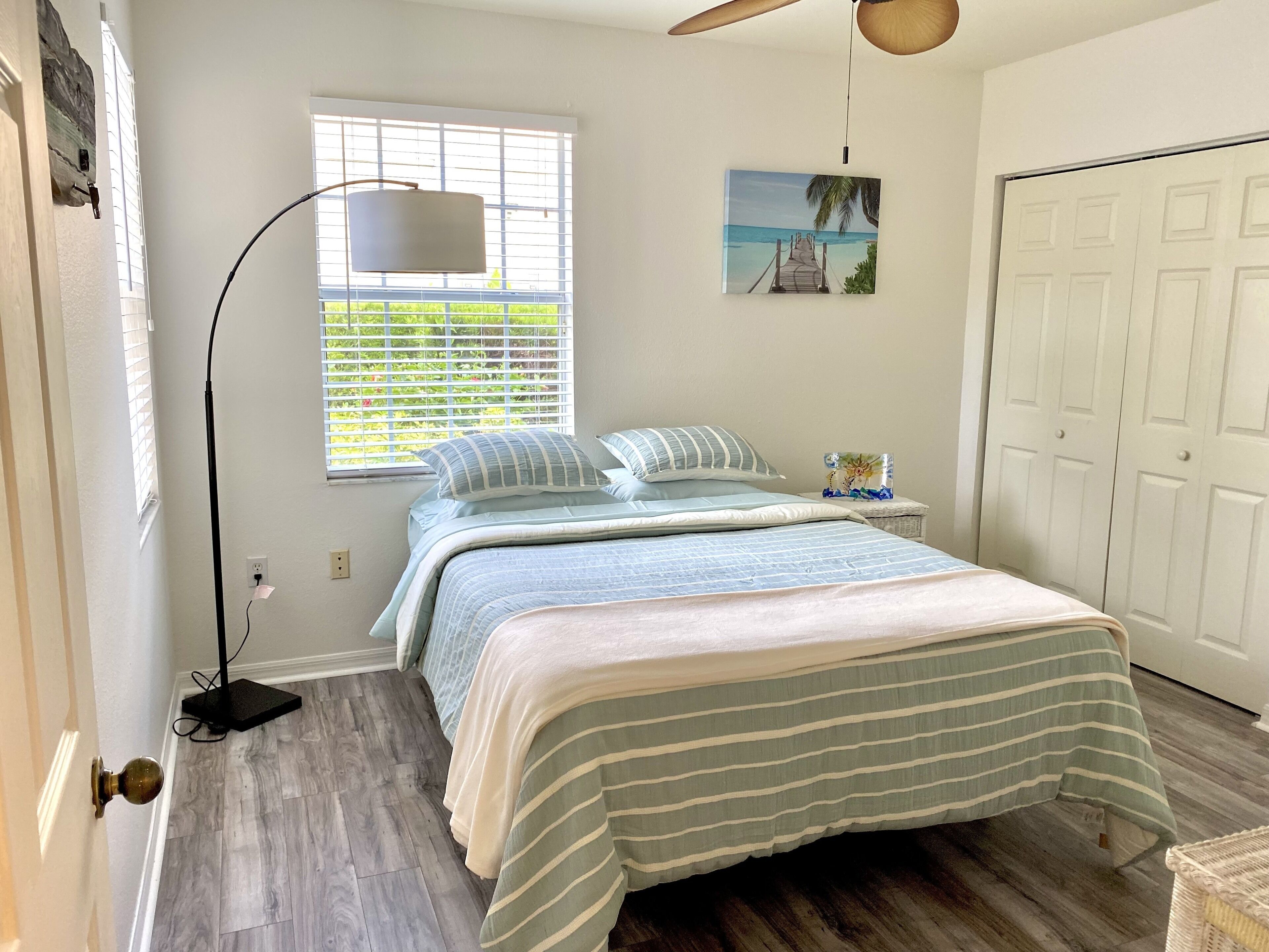 A bedroom with a bed, a window with blinds, a painting on the wall, and a closet.