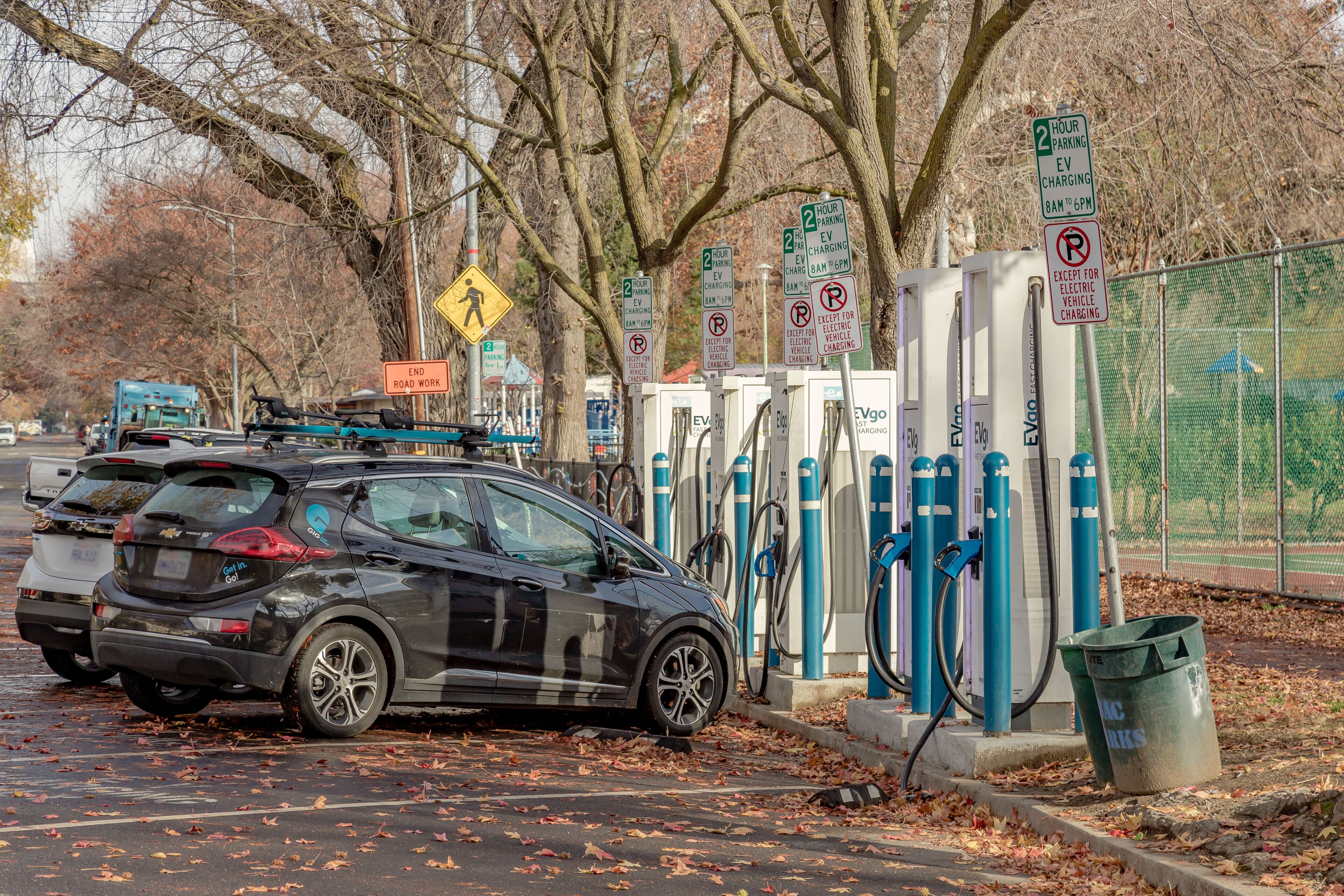 A parking area with electric vehicle charging stations, a pedestrian crossing sign, and a trash bin.