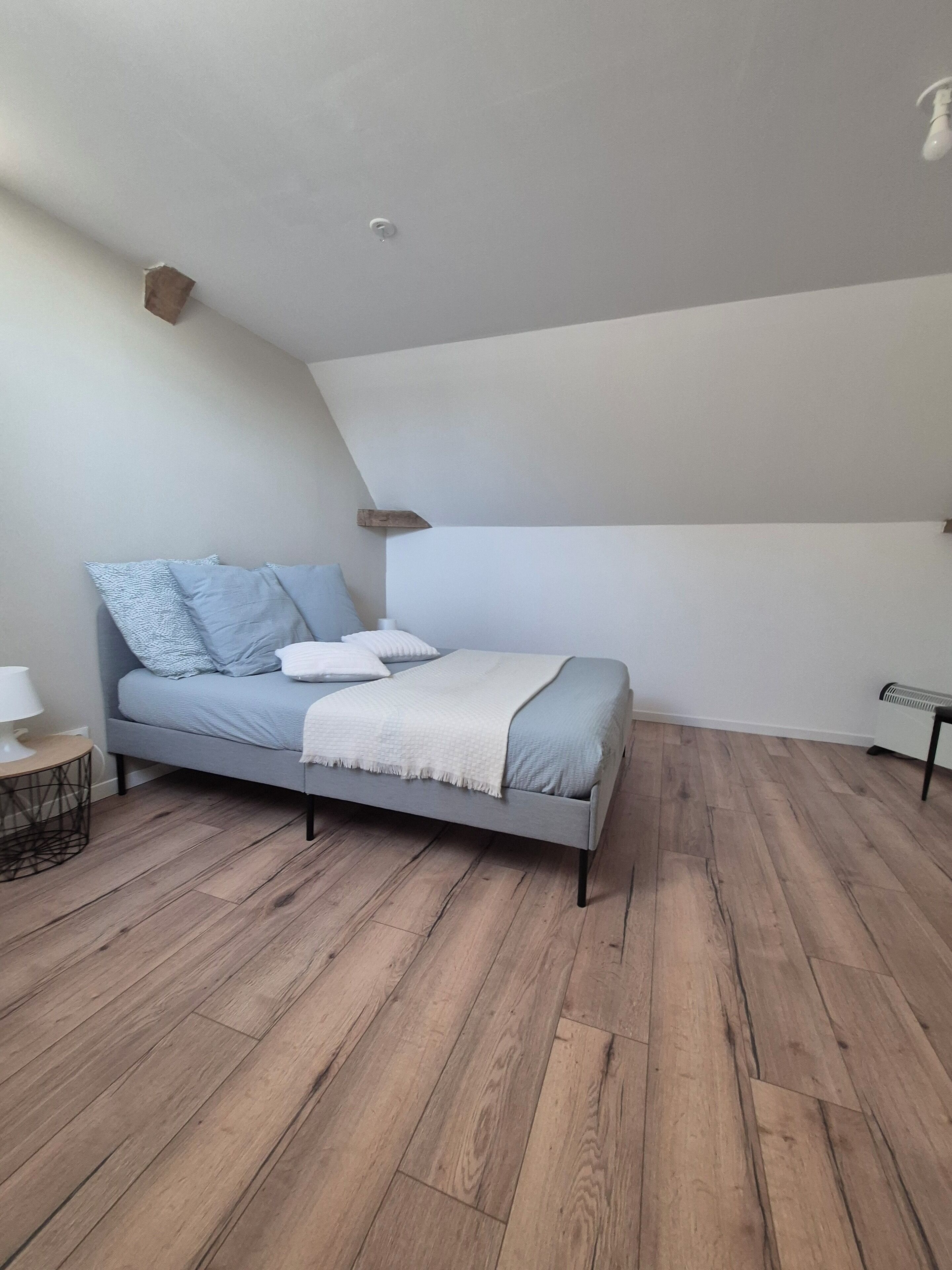 Een moderne slaapkamer met een schuine kap, een grijs bed met wit en blauw beddengoed, een klein nachtkastje met een lamp, en een radiator op de vloer.