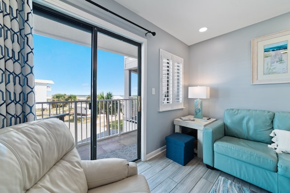A modern living room with a beige sofa, a teal armchair, a small table with a lamp, and a balcony with a view of the outdoors.