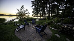 Eine Terrasse mit Adirondack-Stühlen und einem Tisch, mit Blick auf einen See mit einer Anlegestelle und Bäumen im Hintergrund.
