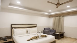 A hotel room with a large bed, a wooden headboard, a ceiling fan, a nightstand, a chair, and a window with curtains.
