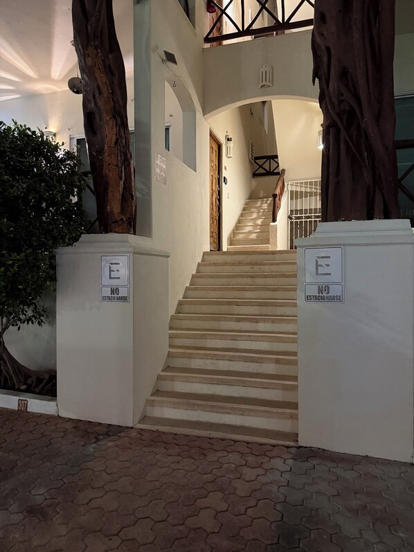 A staircase leading to a door with a 'NO ESTACIONARSE' sign, flanked by large trees and a tiled floor.