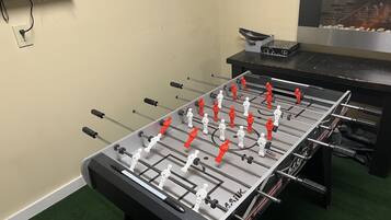 A foosball table in a room with a green carpet, a black table, and a wall-mounted TV.