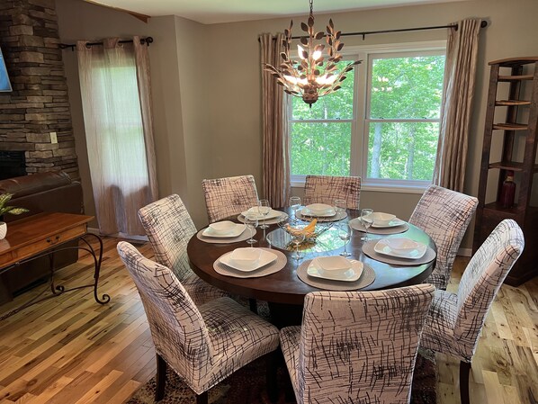 A dining room with a round table set for six, surrounded by patterned chairs, a chandelier, and a view of trees through a window.