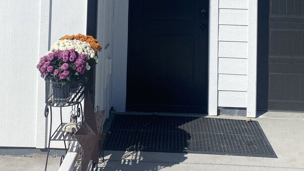 A white building with a black door, a number '105' above the door, a small porch with a wooden beam, and a flower arrangement on the left. - Quiet Country Setting (Bozeman)