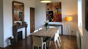 Un espace repas comprenant une table en bois, des chaises blanches, une cheminée, un grand tableau abstrait et un lampadaire.
