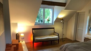 Une chambre avec un plancher en bois, un canapé, une table de chevet, une lampe et une fenêtre donnant sur des arbres.