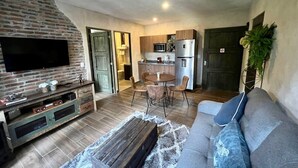 Una sala de estar con televisor, sofá y mesa de centro. Hay una zona de cocina con microondas, refrigerador y mesa de comedor.