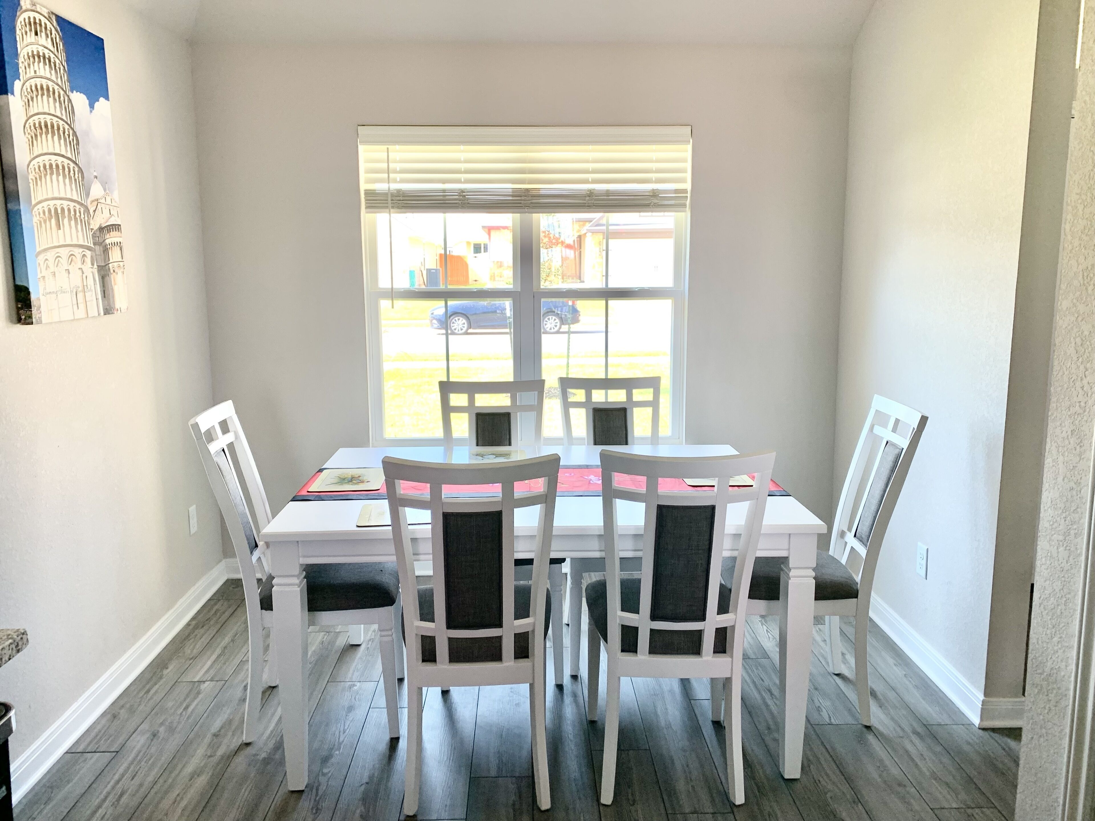 Un comedor con una mesa y sillas blancas, una ventana con persianas, y un cuadro de la Torre Inclinada de Pisa en la pared.