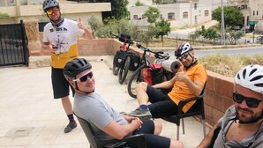 A group of people sitting on chairs outdoors with bicycles nearby, in an urban setting with buildings and trees in the background.