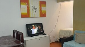 A small, furnished room with a wooden dining table, a TV on a white cabinet, a bed with a blue blanket, and a wall-mounted abstract painting. - Luxury apartment by the sea (Rio de Janeiro)